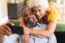 Закрийте щасливу американську пару на пенсії вдома в своїй вітальні, чоловік сидить на дивані з смартфоном, жінка стоїть позаду і обіймає його, дивлячись на телефон разом — стокове фото
