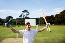 Front view of a teenage Caucasian male cricket player wearing whites, standing on the pitch, smiling and rising his hands, holding a cricket bat and a cricket helmet. — Stock Photo