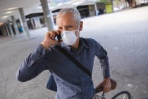 Homme caucasien âgé dehors et autour dans les rues de la ville pendant la journée, portant un masque facial contre le coronavirus, covid 19, assis sur son vélo et en utilisant un smartphone. — Photo de stock