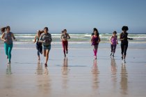 Vue de face d'un groupe multi-ethnique d'amies profitant de l'exercice sur une plage par une journée ensoleillée, courant sur le bord de la mer. — Photo de stock