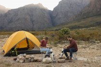 Vue latérale d'un couple caucasien s'amusant lors d'un voyage à la montagne, assis près d'un feu de camp, jouant avec des chiots — Photo de stock