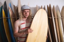 Kaukasischer Surfbrettmacher in seinem Studio, eines der Surfbretter in der Hand und lächelnd in die Kamera, im Hintergrund andere Surfbretter in einem Gestell. — Stockfoto