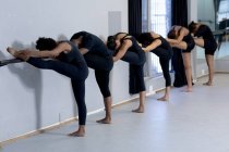 Seitenansicht einer multiethnischen Gruppe fitter männlicher und weiblicher moderner Tänzer in schwarzen Outfits, die während eines Tanzkurses in einem hellen Studio eine Tanzroutine praktizieren, am Geländer stehend und sich streckend. — Stockfoto