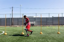 Giocatore di calcio maschile di razza mista con gamba protesica che indossa un allenamento a strisce di squadra in un campo sportivo al sole, riscaldandosi correndo tra i pali. — Foto stock