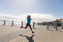 На вигляд спортивна кавказька жінка з довгим темним волоссям, що сидить на прогулянці на березі моря в сонячний день з блакитним небом, біжить. — стокове фото