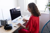 Mulher caucasiana passando tempo em casa, vestindo um vestido rosa, sentada ao lado de sua mesa e usando seu computador. Distanciamento social e auto-isolamento em quarentena . — Fotografia de Stock