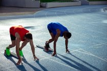 Вид сбоку кавказца и смешанной расы спортсмены мужского пола, практикующие на спортивном стадионе, на позиции на стартовых блоках, готовящиеся к спринту — стоковое фото