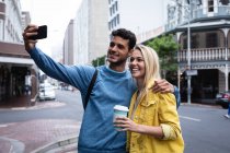 Vorderansicht eines glücklichen kaukasischen Paares, das tagsüber auf den Straßen der Stadt unterwegs ist und sich umarmt, während sie ein Selfie mit ihrem Smartphone machen. — Stockfoto