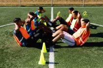 Multi grupo étnico de cinco homens jogadores de futebol um lado vestindo roupas esportivas e coletes de treinamento em um campo de esportes ao sol, aquecendo-se fazendo sentar-se com cones ao lado deles . — Fotografia de Stock