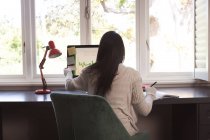 Femme de race mixte passant du temps à la maison, assis au bureau, prenant des notes, travaillant à la maison. Auto-isolement et distanciation sociale en quarantaine pendant l'épidémie de coronavirus covid 19. — Photo de stock