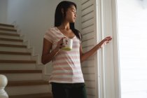 Mujer de raza mixta pasando tiempo en casa, de pie y sosteniendo una taza. Autoaislamiento y distanciamiento social en el bloqueo de cuarentena durante la epidemia de coronavirus covid 19. - foto de stock