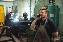 Caucasian male factory worker wearing apron talking on his smartphone with coworker welding in the background. Workers in industry at a factory making hydraulic equipment. — Stock Photo
