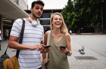 Vista frontal de una feliz pareja caucásica por las calles de la ciudad durante el día, de pie en la calle y usando sus teléfonos inteligentes. - foto de stock
