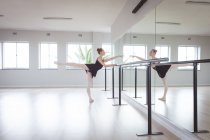 Caucasian attractive female ballet dancer with red hair stretching her leg, preparing for a ballet class in a bright studio, focusing on her exercise, looking at her reflection in the mirror. — Stock Photo