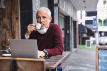 Älterer kaukasischer Mann sitzt an einem Tisch auf einer Kaffeeterrasse, trägt eine Gesichtsmaske gegen Coronavirus, ist 19 Jahre alt, trinkt Kaffee und benutzt einen Laptop. — Stockfoto