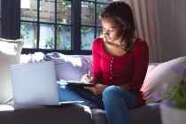 Mulher de raça mista aproveitando seu tempo em casa, distanciamento social e auto-isolamento em quarentena, sentada em um sofá, usando um laptop, escrevendo em seu caderno — Fotografia de Stock