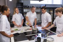 Caucasian group of male and female chefs, listening to a senior Caucasian chef adding ingredients to a pot. Cookery class at a restaurant kitchen. — Stock Photo