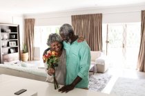 Старшая афроамериканская пара проводит время дома вместе, социальное дистанцирование и самоизоляция в карантинной изоляции во время эпидемии коронавируса 19, обнимая и улыбаясь, женщина, держащая букет цветов — стоковое фото