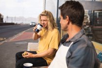 Zwei kaukasische männliche Surfbrettmacher machen während der Arbeit in ihrem Studio eine Pause, sitzen auf einer Bank und interagieren, während sie Kaffee zum Mitnehmen trinken und einen Imbiss einnehmen.. — Stockfoto