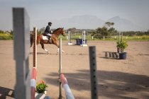 Side view of smartly dressed African American man riding his chestnut horse at a show jumping event on a sunny day, cantering between fences — Stock Photo