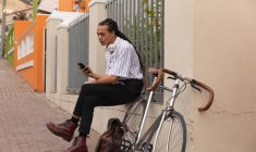 Вид збоку на змішаного гоночного чоловіка з довгими сновидіннями і близько в місті в сонячний день, сидячи на вулиці за допомогою смартфона, з велосипедом, що стоїть поруч з ним . — стокове фото