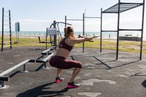 Vue latérale d'une femme sportive caucasienne aux longs cheveux foncés faisant de l'exercice dans une salle de gym extérieure au bord de la mer pendant la journée, faisant des squats. — Photo de stock