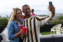 Front view of a Caucasian woman and an African American man hanging out on a roof terrace on a sunny day, taking a selfie and smiling — Stock Photo
