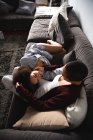 Vista de ángulo alto de una pareja femenina de raza mixta relajándose en casa en la sala de estar en el sofá juntos por la mañana, una mujer acostada, con la cabeza en el regazo de su pareja sentada - foto de stock