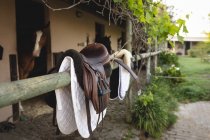 Крупный план коричневого седла с белой седловиной, висящего на деревянном заборе у конюшни в солнечный день, с лошадьми в конюшнях на заднем плане — стоковое фото