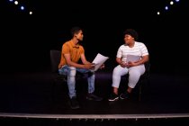 Vue latérale d'un adolescent métis et d'une adolescente afro-américaine du secondaire assise sur des chaises dans un théâtre scolaire vide se préparant avant une représentation, tenant des scénarios et répétant ensemble — Photo de stock