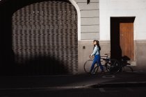 Vue latérale d'une heureuse femme métissée aux cheveux longs et foncés dans les rues de la ville pendant la journée, portant un chapeau, un jean et une veste en denim, marchant avec son vélo avec bâtiment en arrière-plan. — Photo de stock