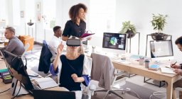 Eine kaukasische Geschäftsfrau arbeitet in einem modernen Büro, trägt ein VR-Headset und berührt einen virtuellen interaktiven Bildschirm, während ihre Kollegen im Hintergrund arbeiten — Stockfoto