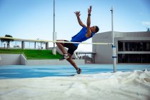 Vue latérale d'un athlète masculin de race mixte pratiquant dans un stade de sport, faisant un saut en hauteur. Entraînement sportif d'athlétisme dans le stade. — Photo de stock