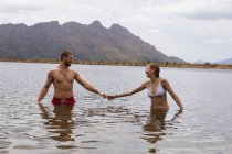 Vista frontale di una coppia caucasica che si diverte durante un viaggio in montagna, in piedi nel lago, tenendosi per mano, guardandosi l'un l'altro — Foto stock