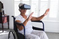 Eine ältere Afroamerikanerin im Ruhestand sitzt an einem sonnigen Tag im Rollstuhl vor einem Fenster und benutzt ein VR-Headset mit ausgestreckten Armen, um sich während der Coronavirus-Pandemie selbst zu isolieren 19 — Stockfoto