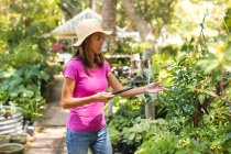 A Caucasian woman wearing a pink t shirt and a straw hat, enjoying time in a sunny garden, touching the leaves of plants and using a tablet computer — Stock Photo