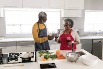 Ein älteres afroamerikanisches Paar, das während der Coronavirus-Epidemie 19 Zeit zu Hause verbringt, soziale Distanzierung und Selbstisolierung in Quarantäne, steht in der Küche und bereitet Essen zu, die Frau hält ein Glas Rotwein — Stockfoto
