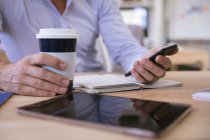 Nahaufnahme eines Geschäftsmannes, der in einem modernen Büro arbeitet, am Schreibtisch sitzt, per Smartphone kommuniziert und Kaffee zum Mitnehmen hält — Stockfoto