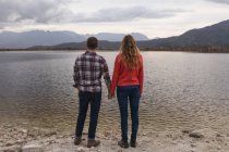 Vista posteriore da vicino di una coppia caucasica che si diverte durante un viaggio in montagna, in piedi su una riva del lago, tenendosi per mano — Foto stock