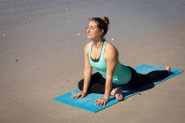 Vista lateral de uma mulher atraente caucasiana, vestindo roupas esportivas, praticando ioga no tapete de ioga, alongando-se na posição de ioga, na praia ensolarada. — Fotografia de Stock