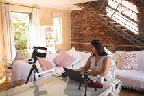 Mulher branca vlogger em casa, em sua sala de estar usando uma câmera e um laptop para preparar seu blog online. Distanciamento social e auto-isolamento em quarentena . — Fotografia de Stock