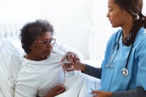 Старшая женщина смешанной расы проводит время дома, лежа в постели, к ней приходит медсестра смешанной расы, медсестра дает ей стакан воды — стоковое фото