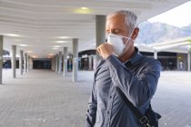 Homem caucasiano sênior nas ruas da cidade durante o dia, usando uma máscara facial contra o coronavírus, vívido 19, cobrindo seu rosto enquanto tosse . — Fotografia de Stock