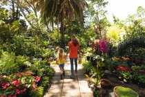 Una donna caucasica e sua figlia si godono il tempo insieme in un giardino soleggiato, tenendosi per mano e camminando lungo un sentiero tra le piante — Foto stock