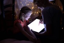 Вид сбоку на кавказку, наслаждающуюся семейным отдыхом с дочерью дома вместе, сидящую в палатке в своей гостиной, улыбающуюся и пользующуюся цифровым планшетом — стоковое фото