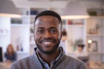 Портрет щасливого афроамериканського бізнесмена, який працює в сучасному офісі, дивиться на камеру і посміхається, зі своїми колегами, які працюють на задньому плані — стокове фото