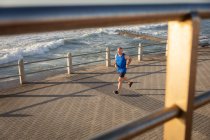 Vista lateral de ángulo alto de un hombre mayor caucásico maduro haciendo ejercicio en un paseo marítimo en un día soleado, corriendo - foto de stock