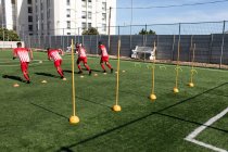 Multi equipe étnica do sexo masculino cinco jogadores de futebol um lado vestindo um treinamento de tira de equipe em um campo de esportes ao sol, aquecendo-se correndo . — Fotografia de Stock