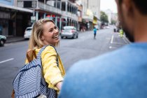 Visão traseira de uma mulher caucasiana feliz com longos cabelos loiros, andando pela rua, segurando uma mão de seu parceiro, sorrindo . — Fotografia de Stock