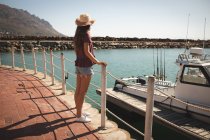 Una adolescente caucásica, con sombrero de paja, disfrutando de su tiempo en un paseo marítimo, en un día soleado, apoyada en una barrera, mirando hacia otro lado - foto de stock